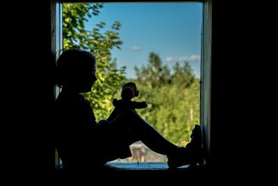 Silhouette man sitting by window against sky