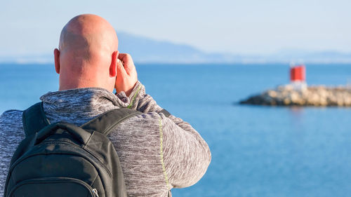 Rear view of a man with shaved head hair, taking photos of a seascape in the port.