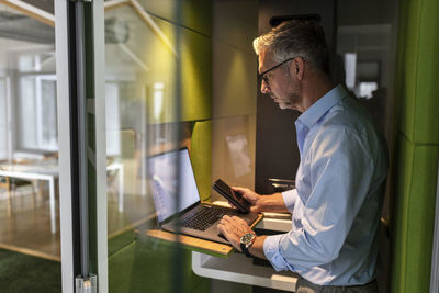 Mature businessman holding mobile phone while using laptop in soundproof cabin at office