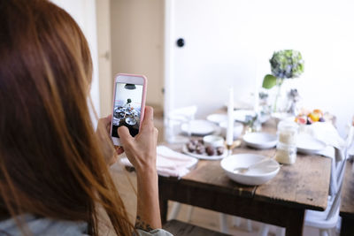 Rear view of woman photographing plates and food on table by wall at home