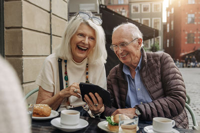 Cheerful senior couple sharing smart phone while sitting at sidewalk cafe