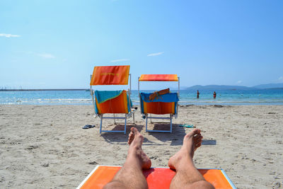 Low section of man resting on lounge chair at beach against blue sky