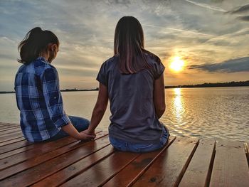 Rear view of women sitting on shore against sky during sunset