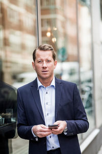 Contemplating mature businessman with smart phone looking away while standing by glass window