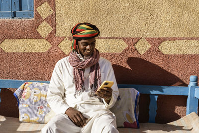 Cheerful egyptian male wearing traditional turban and clothes surfing internet on smartphone while sitting on sofa in courtyard near house on sunny day