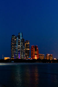 Illuminated buildings by sea against clear blue sky at dusk