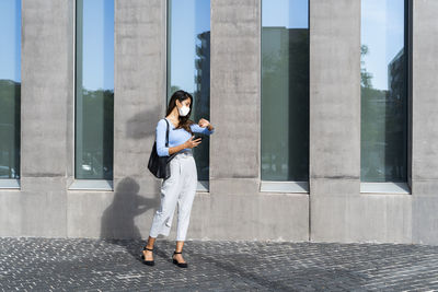 Businesswoman in face mask checking time on wristwatch while standing on footpath during pandemic