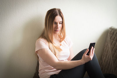 Woman using phone while sitting against wall at home