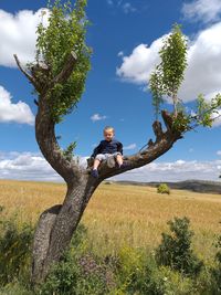 Young man sitting on field by tree against sky