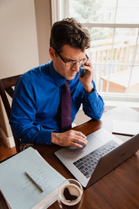 Man on cellphone working from home using a computer at a dining table.