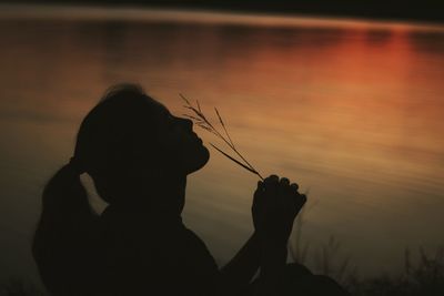 Silhouette young woman holding plant while sitting by lake during sunset