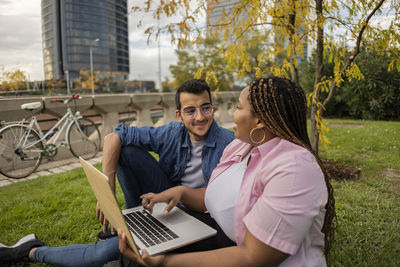 Smiling man and woman discussing and sitting with laptop on grass