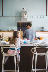 Rear view of girl looking away while sitting at kitchen island with father standing in background