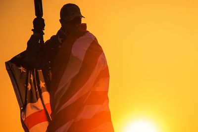 Soldier wrapped in american flag standing against sky during sunset