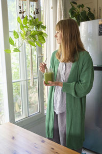 Blond woman with green smoothie looking out through window