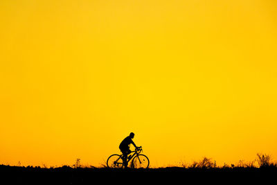 Silhouette man cycling on field against orange sky