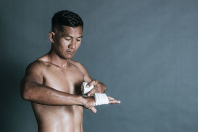 Shirtless man wrapping bandage on hand against wall