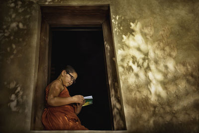 Side view of boy reading book while sitting on window