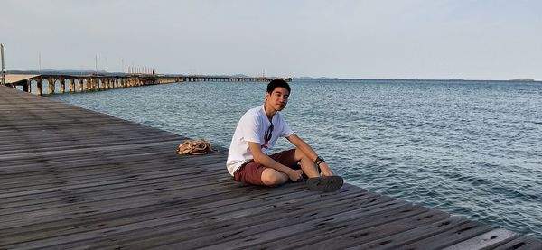 Portrait of man sitting on pier by sea against clear sky