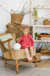 Three-year-old girl in a dress barefoot sits on a chair with a white rabbit in the living room