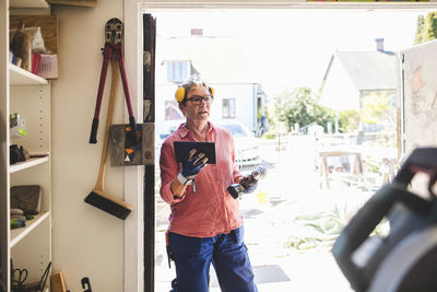 Senior woman holding drill and digital tablet while standing at doorway of workshop