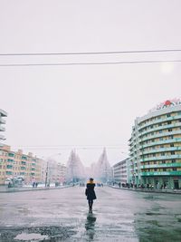 Rear view of woman standing on wet road amidst buildings against sky