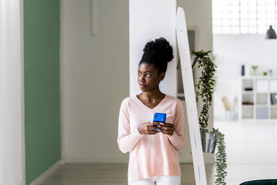Thoughtful woman with mobile phone looking away while standing at home