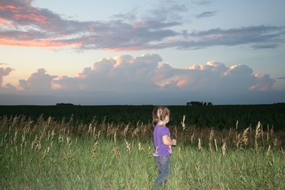 Side view of girl standing on grassy field against sky during sunset