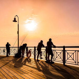 Silhouette men on footbridge over sea against orange sky