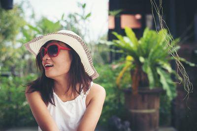 Smiling young woman wearing sunglasses and hat while sitting at yard