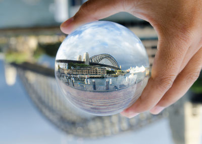 Cropped hand holding crystal ball with reflecting of bridge against cloudy sky