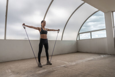 Determined young ethnic female athlete in sportswear working out with elastic band while looking forward