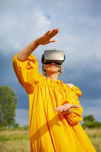 Woman gesturing while wearing vr glasses standing against sky