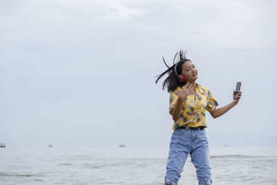 Girl dancing on music with mobile phone and headphones while standing on beach against sky