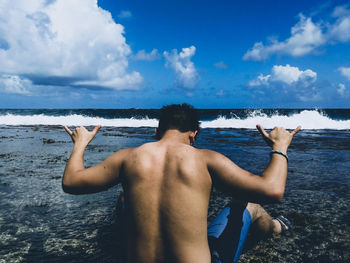 Rear view of shirtless man gesturing while sitting at beach against sky