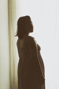 Young pregnant woman standing by window at home