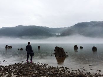 Full length rear view of woman standing by river against mountains and sky during foggy weather