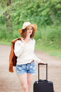 Portrait of woman with luggage wearing hat standing on road