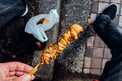 Low section of man holding yakitori while sitting at footpath