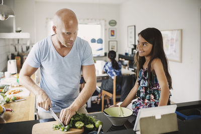 Smiling daughter looking at father chopping vegetables at kitchen counter