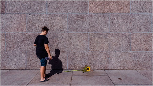 Rear view of boy standing by sunflower on footpath against wall during sunny day