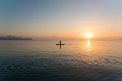 Back view of silhouette of unrecognizable female surfer standing on paddleboard and rowing against spectacular sun in sunset sky