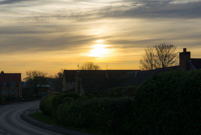 Road by buildings against sky during sunset