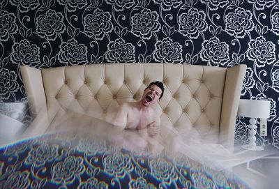 Double exposure of shirtless man sitting on bed against floral wallpaper at home