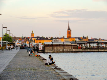 People sitting on bridge over river in city against sky