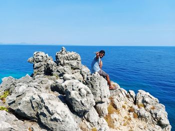 Man standing on rocks by sea against blue sky