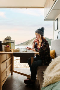 Side view of focused female traveler creating handmade accessories while sitting at wooden table in parked truck at seaside