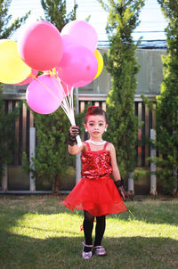 Portrait of cute girl holding balloons while standing on grassy field against fence