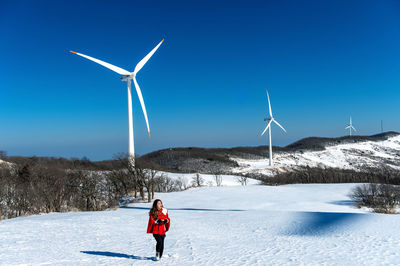 Full length of woman walking on snow covered road against wind turbines and clear blue sky