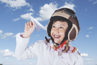 Close-up of happy boy holding paper airplane while wearing helmet against cloudy sky